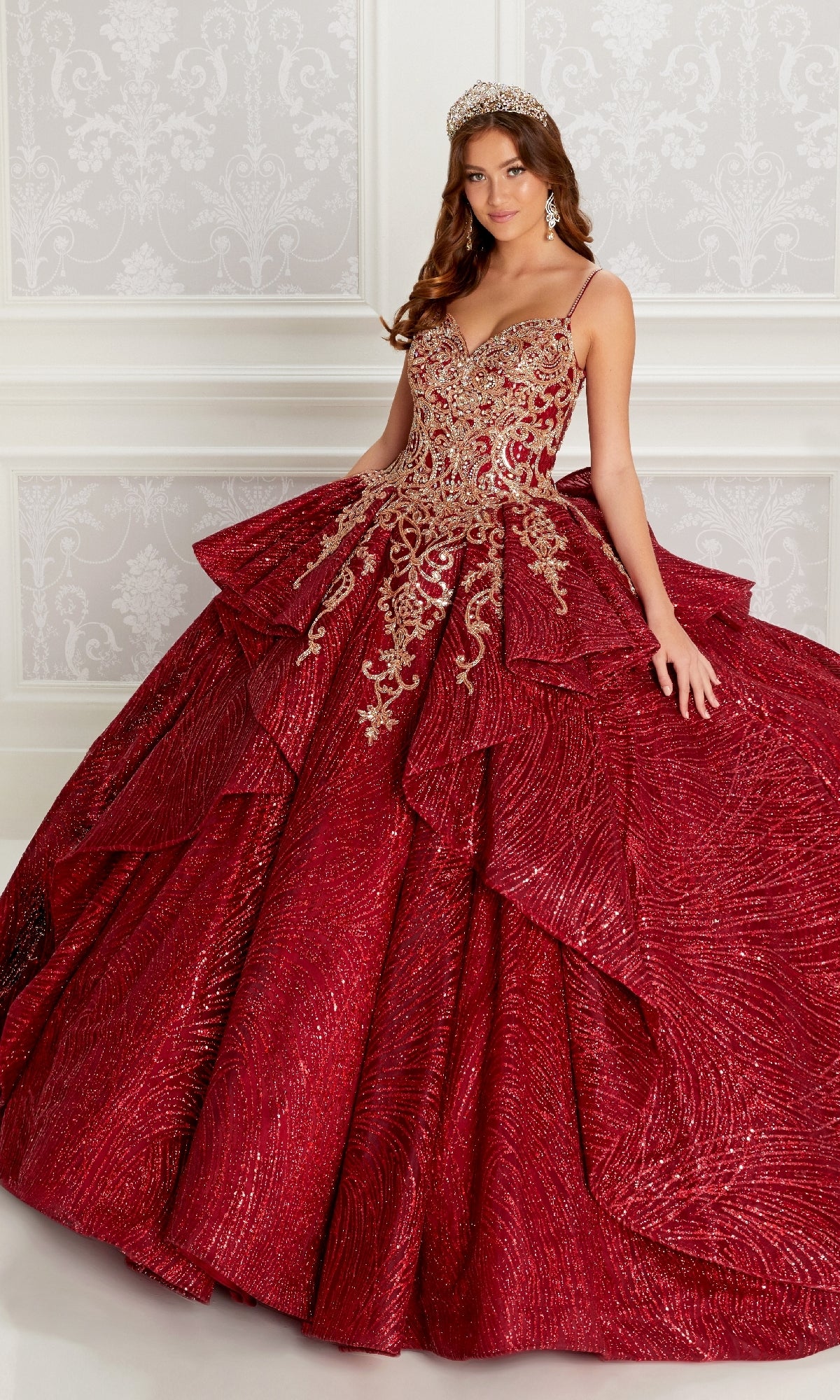 A woman in the Quinceanera Dress PR22142 By Princesa, featuring a sparkling red design with gold embroidery, corset back, detachable sleeves, and a tiara, stands before an ornate white wall radiating elegance.